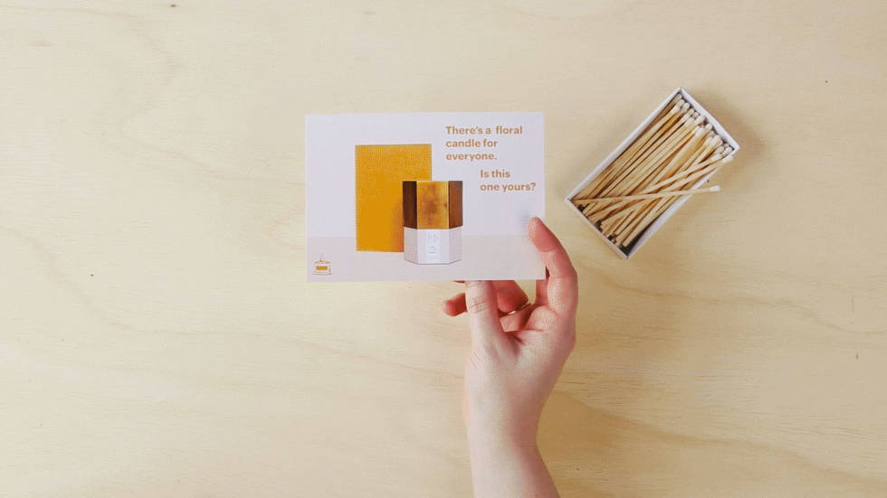 A photo of a person's hand turning a postcard back and forth to show how the copy on both sides works together. Copy on front: "There's a floral candle for everyone. Is this one yours?" Copy on back: "Get your free candle" 