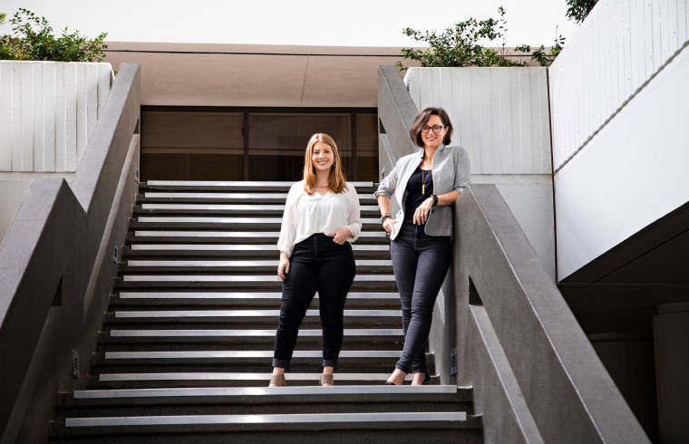 Mary Frances and Jamie of the Roadie team standing together on steps outside. Both women are smiling and looking downward at the photographer. 